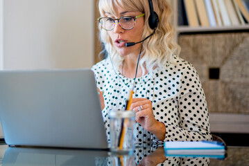 Wall Mural - Busy adult woman at work at laptop computer calling on headphones and microphones. Concept of modern online job female people. Attractive lady with eyewear in communication