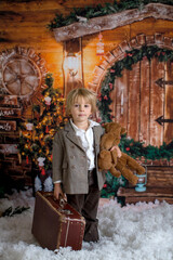 Wall Mural - Cute fashion toddler boy, playing in the snow with teddy bear in front of a wooden cabin log