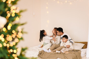 A mixed race Asian family with two children in cozy sweaters is relaxing together, resting and having fun on a bed in a decorated bedroom with Christmas tree at home in the New Year. Selective focus