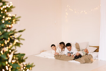 A mixed race Asian family with two children in cozy sweaters is relaxing together, resting and having fun on a bed in a decorated bedroom with Christmas tree at home in the New Year. Selective focus