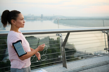 Confident motivated sporty woman wearing in pink t-shirt, and smartphone holder on her hand holding earphones and looking away getting ready for morning jog at dawn on a treadmill of a city bridge