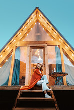 Woman Is Sitting On The Porch Of A Wooden Hut With Lights Of Garlands In The Evening. The Concept Of Glamping And Renting A Chalet.