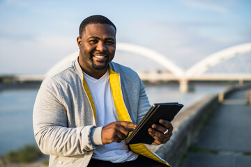 Modern black man using digital tablet while sitting by a river.