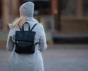 Wall Mural - Happy young blonde teenage girl going to school or college wearing black backpack. Blonde girl in rgay hat and gray coat. Pictuce from back