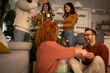 Wall Mural - Young couple in love sitting on a sofa next to a Christmas tree. The guy gives the woman a gift for the new year