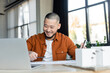 smiling asian architect holding smartphone while working with blueprints near blurred laptop