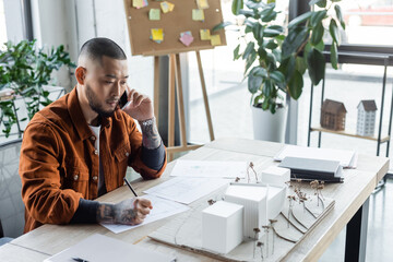 Sticker - asian architect talking on mobile phone near blueprints and house models on work desk