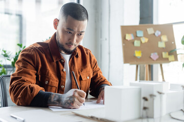 Sticker - tattooed asian architect looking at blurred house models while drawing blueprint in office