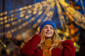 Wall Mural - Christmas, New Year, winter holiday conception: happy smiling woman wearing knitted beanie hat, gloves, faux fur coat, posing in street. Copy, empty space for text