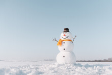 Funny Snowman In Stylish Brown Hat And Yellow Scalf On Snowy Field. Blue Sky On Background