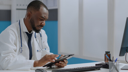 Wall Mural - African american therapist doctor holding tablet computer checking sickness report typing medical expertise. Physician man working at healthcare treatment in hospital office. Medicine concept