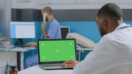 Wall Mural - African american doctor looking at mock up green screen chroma key laptop computer with isolated display. Therapist man analyzing sickness diagnostic working at medical report in hospital office