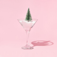 Christmas tree and snow in martini cocktail glass on pastel pink background. Winter creative idea. Retro aesthetic christmas minimal concept.