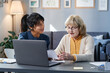 © AnnaStills - Senior woman studying at the table with laptop, and volunteer helping her in her domestic study