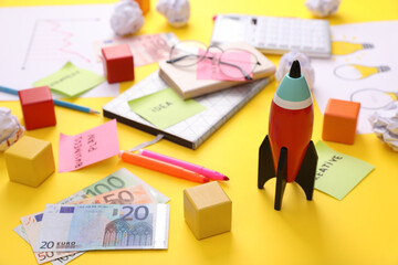 Poster - Composition with toy rocket, stationery and money on yellow background. Startup concept