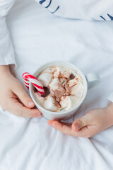 Adorable 6 year old boy in festive pajamas sits by the Christmas tree holding a mug of hot cocoa with marshmallows and lollipop in Christmas decorations. Winter holidays. New Year. Cozy Home.