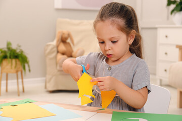 Little girl cutting color paper with scissors at table indoors