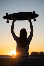 Freedom And Urban Fashion: Teenage Girl With Longboard Looking At Sunset, Evening Sky On Background. Silhouette Female Longboarder Teen Or Student Enjoy Summer. Happy Casual Woman With Skateboard
