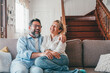 © Daniel - Happy caucasian couple laughing while sitting on sofa in the living room. Husband and wife laughing out loud sitting on couch at home. Man and woman spending leisure time at modern apartment