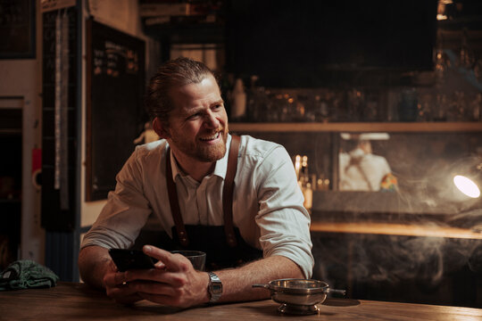 Caucasian male bartender sitting at bar texting on cellular device laughing 