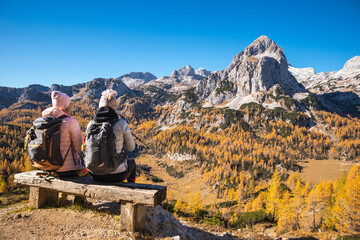 Two female hikers on a bench enjoying mountain panorama view on a sunny autumn day with golden larch trees.