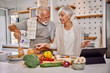 © kerkezz - Smiling elderly vegan couple preparing a fresh vegetable salad in the kitchen, using a tablet