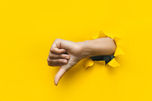Hand Showing A Thumb Down Through Ripped Hole In Bright Yellow Paper Background. Concept Of Dislike And Disapproval Gesture.