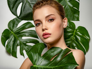 Wall Mural - Closeup face of young beautiful woman with a healthy clean skin. Beautiful white girl with big green leaves. Beauty and spa treatment concept. Pretty woman with natural makeup and plant near face