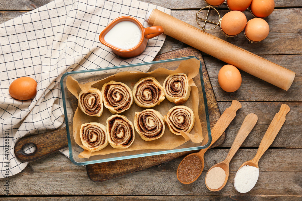 Baking dish of uncooked cinnamon rolls and ingredients on wooden background