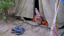 Bare Feet Male Legs Peeking Out From An Open Military Tent In Nature. Tourist Is Resting, Sleeping At Camping, Airing Naked Legs. Traveler Relaxes In A Tent Without Slippers Among Trees Outdoors. 4K.