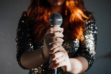 Faceless Portrait Of Redhead Woman In Sparkly Evening Dress Holding Microphone On Dark Night Background. Unrecognizable Female Singer With Modern Microphone In Hands
