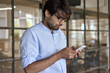 © insta_photos - Young Indian business man using cellphone apps standing in office. Arab professional businessman holding mobile phone managing finance banking applications corporate digital technology.