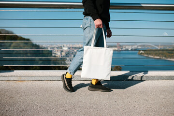 Cropped photo of a young man with a white fabric eco bag against an urban city. Eco friendly. Reusable bag in young man's hand. Mockup for print