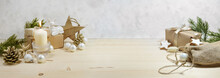 Natural Christmas Decoration With A Warm White Candle, Baubles, Stars, And Gifts On A Wooden Table Against A Light Plastered Wall, Panoramic Format, Copy Space, Selected Focus, Narrow Depth Of Field