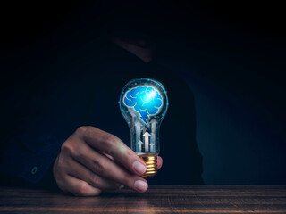 Poster - Creative, idea, innovation, knowledge, and inspiration concept. The human brain icon with rising arrows glowing inside the digital light bulb in man's hand on the wooden table and dark background.