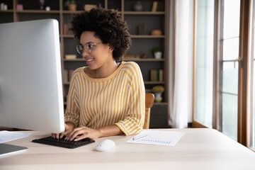 Happy young Black student girl in glasses working at desktop, using big monitor, typing on wireless keyboard. Remote worker, designer, professional developer doing online tasks at workplace.