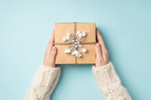 First Person Top View Photo Of Female Hands In White Sweater Holding Craft Paper Giftbox Decorated With Snow Twig And Twine On Isolated Pastel Blue Background