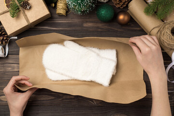 Wall Mural - First person top view photo of young woman's hands packing white fluffy mittens in kraft paper green gold christmas tree balls pine twigs and handicraft tools on isolated wooden desk background