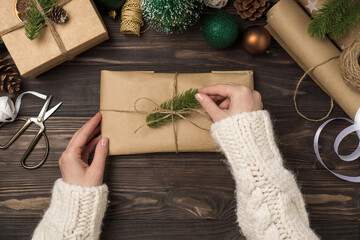 Wall Mural - First person top view photo of female hands in knitted pullover decorating craft paper giftbox with pine twig and twine handicraft tools cones green gold balls on isolated wooden desk background