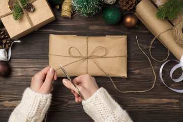 Wall Mural - First person top view photo of female hands in sweater packing kraft paper giftbox with twine bow scissors green gold balls pine twigs and handicraft tools on isolated dark wooden desk background