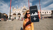 Audio guide online app on digital mobile smartphone. Happy young student woman holding phone listening audioguide at San Marco square in Venice, Italy. Devices for listening to excursions.