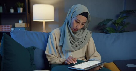 Wall Mural - Attractive cheerful young Asian muslim beauty woman in hijab casual sitting on couch with laptop use digital tablet work on cozy living room in home at night. Girl people Islam lifestyle concept.