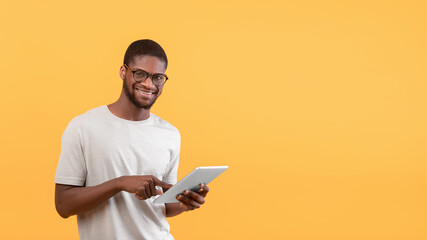 Wall Mural - Portrait of happy black man using digital tablet and smiling to camera, standing over yellow background, copy space