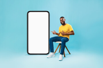 Poster - Happy black guy sitting on chair, pointing at giant smartphone with white screen, offering mockup for mobile app design