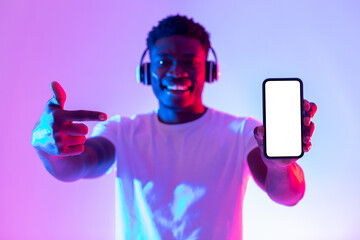 Poster - Check out new music app. Happy black guy in headphones pointing at smartphone with empty screen in neon light, mockup
