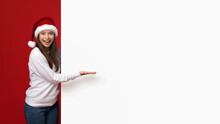 Happy Excited Young Woman Wearing Santa Hat Pointing At White Advertisement Board