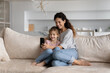 © fizkes - Happy family mother and little daughter having fun with modern smartphone, sitting on couch at home, smiling mom with cute girl kid chatting online by video call, taking selfie, watching cartoons