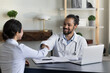 © fizkes - Smiling African American man doctor in white uniform with stethoscope and Indian patient client shaking hands, making medical insurance agreement, sitting at desk in office, healthcare concept