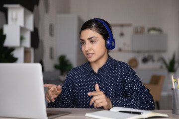 Canvas Print - Concentrated young Indian business woman in headphones communicating distantly with colleagues or clients by video call conversation, discussing working issues or project ideas remotely.