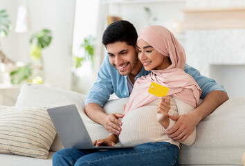 Wall Mural - Purchasing goods online. Happy muslim pregnant couple using laptop computer and credit card, sitting on sofa